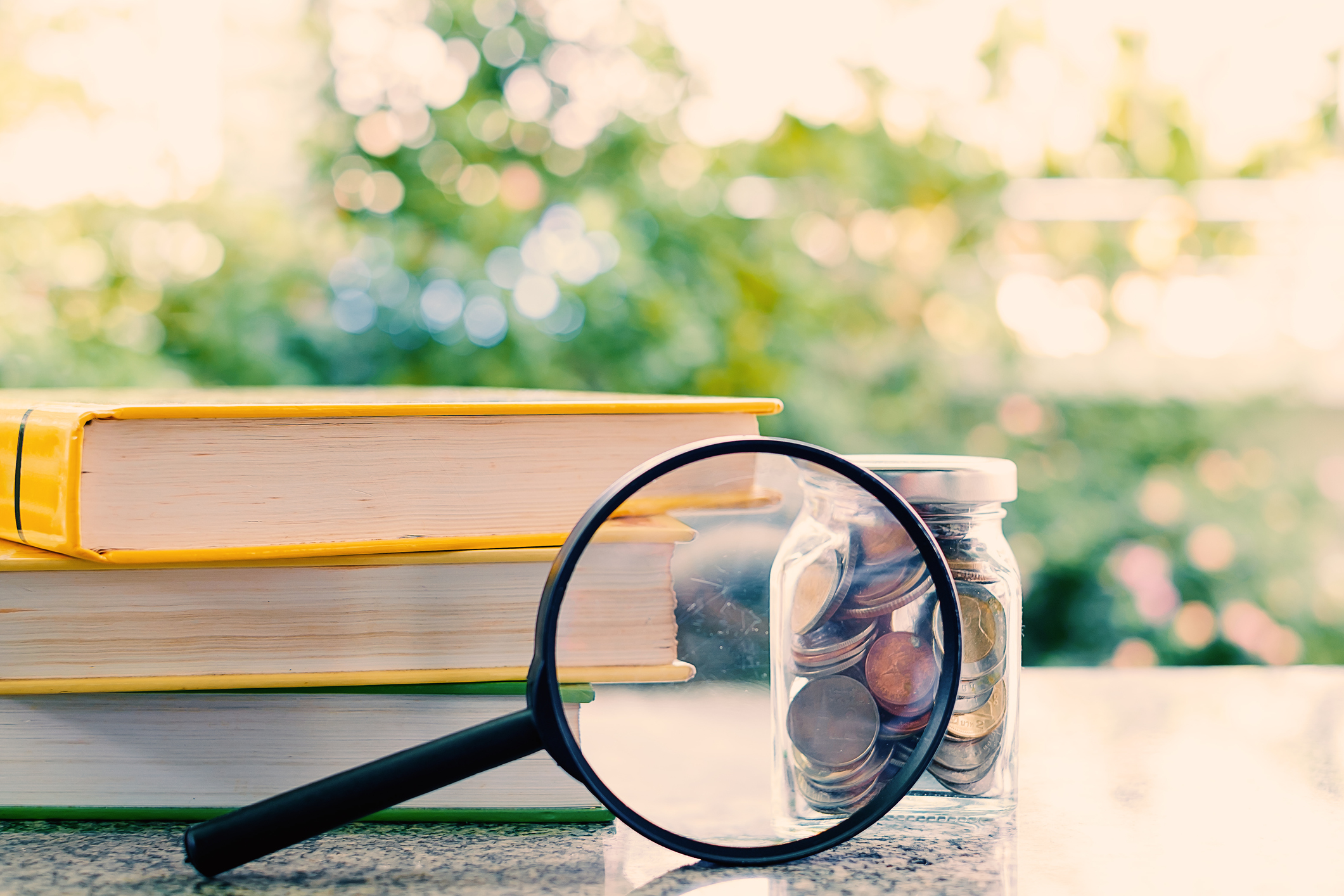 Books and money coins in the glass jar zoomed by the magnifying glass on blurred natural green background and added colour filter for financial and education concept Books and money coins in the glass jar zoomed by the magnifying glass on blurred natural green background
